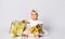 Baby girl in dress and headband, barefoot. She holding golden gift box, sitting on floor isolated on white. Close up, copy space