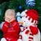 Baby girl toddler in red dress sitting by New Year tree with snowman toy closing covering her eyes with hands