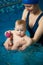 Baby holding toy balls and sitting in his mother`s hands in clear blue water of swimming pool during wellness treatments