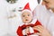 A baby in a little Santa suit in dad`s arms. The father looks at him and enjoys his fatherhood and is ready to celebrate Christma