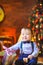 Baby sits on the floor with a small gift in hand and smiles and grimaces, on the background of bright festive lights