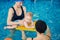 Baby in swimming nappies lying down on floating board. Master carrying out swimming courses for moms and babies in pool