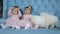 Babyhood, little female twins in pink dresses and with bows on head posing for photo sitting on sofa with white puppies