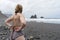 Back Blond curvy lady putting bikini, standing in a black sand beach, rocky. Cloudy day and girl looking the sea horizon in