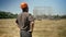 Back of confident builder looking at metal construction on background, standing in helmet on straw field