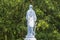 The back of a female Statue of Hope in a graveyard against green trees