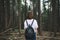 Back of a hipster girl with a backpack stands in a dark dense forest and looks forward. Stylish hiker girl in casual clothes