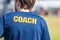 Back of male sport coach wearing blue COACH shirt at an outdoor sport field