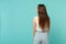 Back rear view of attractive young woman in light casual clothes looking aside isolated on blue turquoise wall