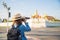 Back side Asian woman in jeans jacket and straw hat carrying a mini backpack taking a photo in front of Wat Phra Kaew one of