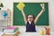 Back to school. Little happy schoolchild raised hands up against the background of a green blackboard in a classroom.