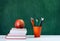 Back to school, orange pencil holder, stack of books on white table with red apple, empty green school board background, education