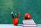 Back to school, orange pencil holder, stack of books on white table with red apple, empty green school board background, education