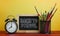 Back to School Text. Alarm Clock, Blackboard and School Stationary in Basket on Wooden Table Yellow Background