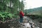 Back of tourist woman in red raincoat stands in forest on rocks in mountain stream, looking forward. Girl walking in mountainous