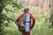 Back view of active backpacker looking for way out in forest, having sleeping pad and dark rucksack with thermos at his back,