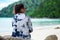 back view. asia woman wearing sunglasses and indigo shirt sitting on rock. front of her are sea and green forest. this image for