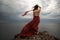 Back view a beautiful sensual woman in a red long dress poses on a rock high above the sea during sunset. Dark clouds all over the