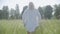 Back view of blond Caucasian overweight woman walking in sunrays on green summer meadow. Middle shot of plump young lady