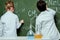 Back view of boy and girl in lab coats drawing chemical formulas on chalkboard