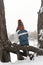 Back view of boy in knitted hat and overalls sits on a tree branch in the snow-struck forest. Winter holidays. Outdoors winter