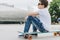 Back view. Boy in white T-shirt and blue jeans, sits on city street on skateboard. Space for text, logo, image. Mock up.