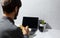 Back view of businessman working home, making notes in notebook, using wireless earphones, laptop, coffee mug and cactus on desk.