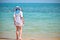 Back view of child girl in white dress and big hat with blue ribbons in long braids standing barefooted on beach