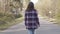 Back view of confident Caucasian brunette woman walking along empty road in sunny forest. Millennial female tourist