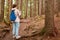 Back view of experienced traveller standing in middle of forest, getting lost, looking for way out, using her smartphone, taking