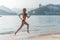 Back view of fit slim girl running barefoot on seashore wearing bikini. Young woman doing cardio exercise beach lit in