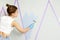 Back view of girl painting wall with paint roller and using masking tape while standing on ladder