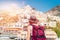 Back view of a girl in a white dress, a hat stands on the beach in Positano. View of houses and hotels in the background. Travel