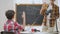 Back view of interested schoolboy raising hand in classroom and asking question with blurred teacher standing at