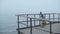 Back view of little girl in rubber boots and plaid standing on wooden pier near the sea. Alone child in foggy windy day.