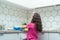 Back view of little preteen girl with loose long dark hair wearing taking off blue rubber gloves, standing near table.