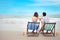Back view of in love asian couple who wearing swimming suit sitting on deck chair at tropical beach with orange and beautiful blue