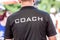 Back view of a male coach wearing black coach shirt with the white word COACH printed on back
