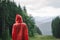 Back view of a male tourist in a bright red raincoat wearing a hood on a rainy day in the mountain fir woods on the hill. Copy