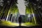 Back view of man with head flashlight standing on forest ground road among tall brightly illuminated spruce trees under beautiful