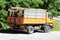 Back view of old yellow truck filled with firewood parked on gravel parking lot next to paved road surrounded with dense trees