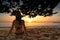 Back view of pregnant woman sit on sand and watching sunset at tropical beach. Woman wear swimsuit and straw hat relaxing