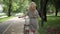 Back view of relaxed Caucasian middle aged woman in grey dress walking with bike in summer park outdoors. Camera follows