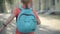 Back view of schoolboy with backpack running along sunny schoolyard outdoors. Blond positive Caucasian boy hurrying for