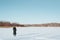 Back view of single woman tourist with backpack standing on sunny frosty day on frozen lake against backdrop of forest