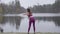 Back view of slim Caucasian brunette girl stretching on lake shore. Workout of confident slender woman outdoors. Sport