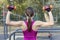 The back view of slim girl in bright sportswear training with dumbbells for biceps at outdoor sportground. White earphones, protec