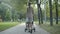 Back view of slim stylish woman in elegant dress and high-heels walking with baby stroller in summer park. Wide shot of