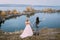 Back view of stylish couple newlyweds with hands up are posing before a lake on the hill. Autumn wedding ceremony