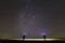 Back view of two men with head flashlights on green grassy field under dark blue summer starry sky.